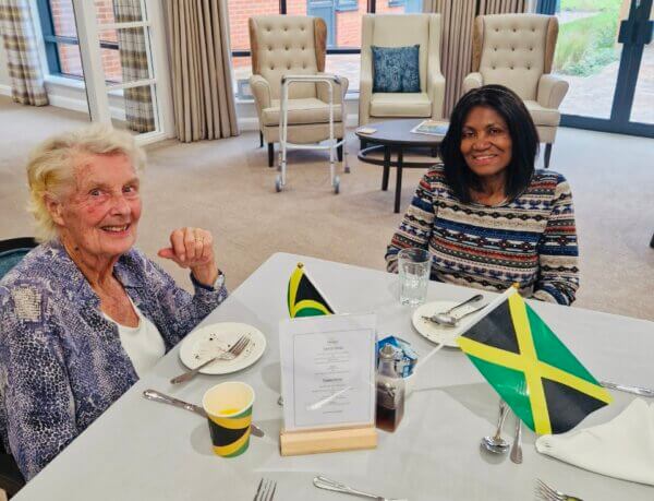 Two elderly women sitting at a dining table decorated with Jamaican flags, smiling at a care home event.